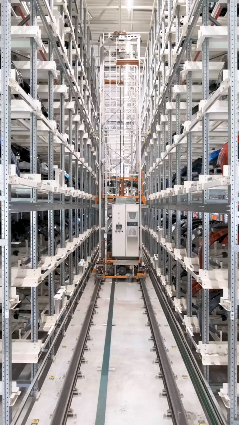 Automated retrieval robot moves along the rails that line the walkway between the industrial shelving racks that store car parts.